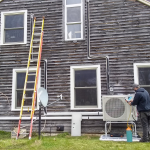 An air-source heat pump outdoor unit being installed outside a brown house